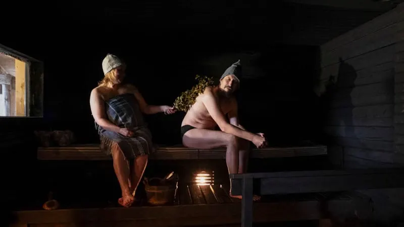 Interior escuro da Seven Star Smoke Sauna do Isokenkäisten Klubi, com duas pessoas de gorro de sauna no banco e uma vihta de bétula